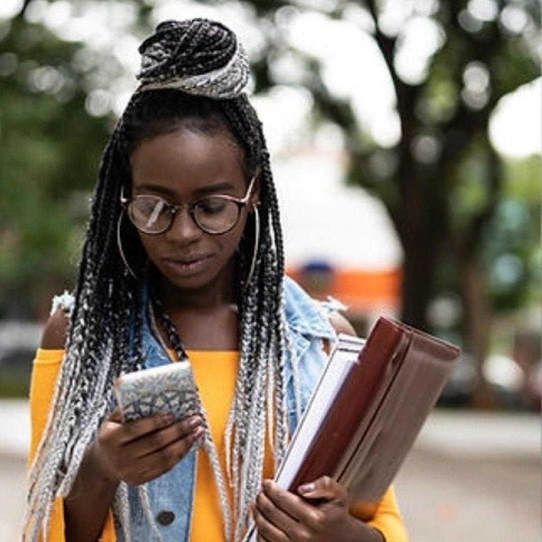 A picture of a woman with glasses looking down at her phone in one hand while the other hand is holding onto books.
